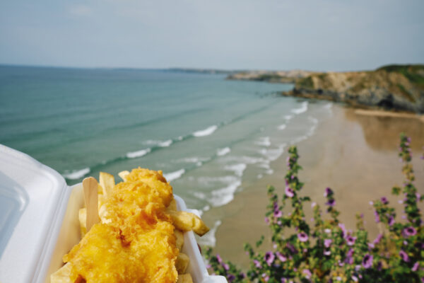POV of a hand holding a carton of Fish and Chips at the Beach, Newquay, Cornwall on a sunny day.